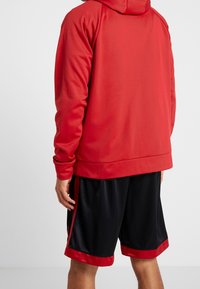 Sudadera roja con capucha de textura suave y mangas largas, combinada con pantalones cortos deportivos negros con ribetes rojos y detalles laterales.