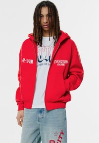 Rote Reißverschluss-Hoodie aus Fleece, mit weißen Texten und Grafiken. Getragen über einem weißen T-Shirt und kombiniert mit blauen Jeans.