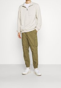 Sweat-shirt beige à fermeture éclair avec une poche poitrine, associé à un pantalon cargo vert olive avec poches latérales et poignets élastiques, porté avec des baskets blanches.