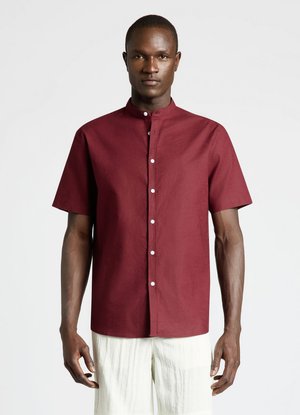 Homme portant une chemise bordeaux à manches courtes boutonnée avec un col mandarin et un pantalon texturé blanc cassé sur fond uni.