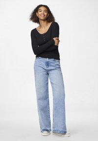 Black long-sleeve ribbed top with button detailing, paired with light blue wide-leg jeans and white sneakers, against a neutral background.