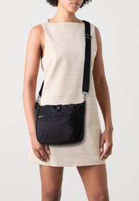 Černá nylonová crossbody taška s přední kapsou, stříbrným kováním a nastavitelným popruhem, vystavená na světlých kostkovaných šatech.