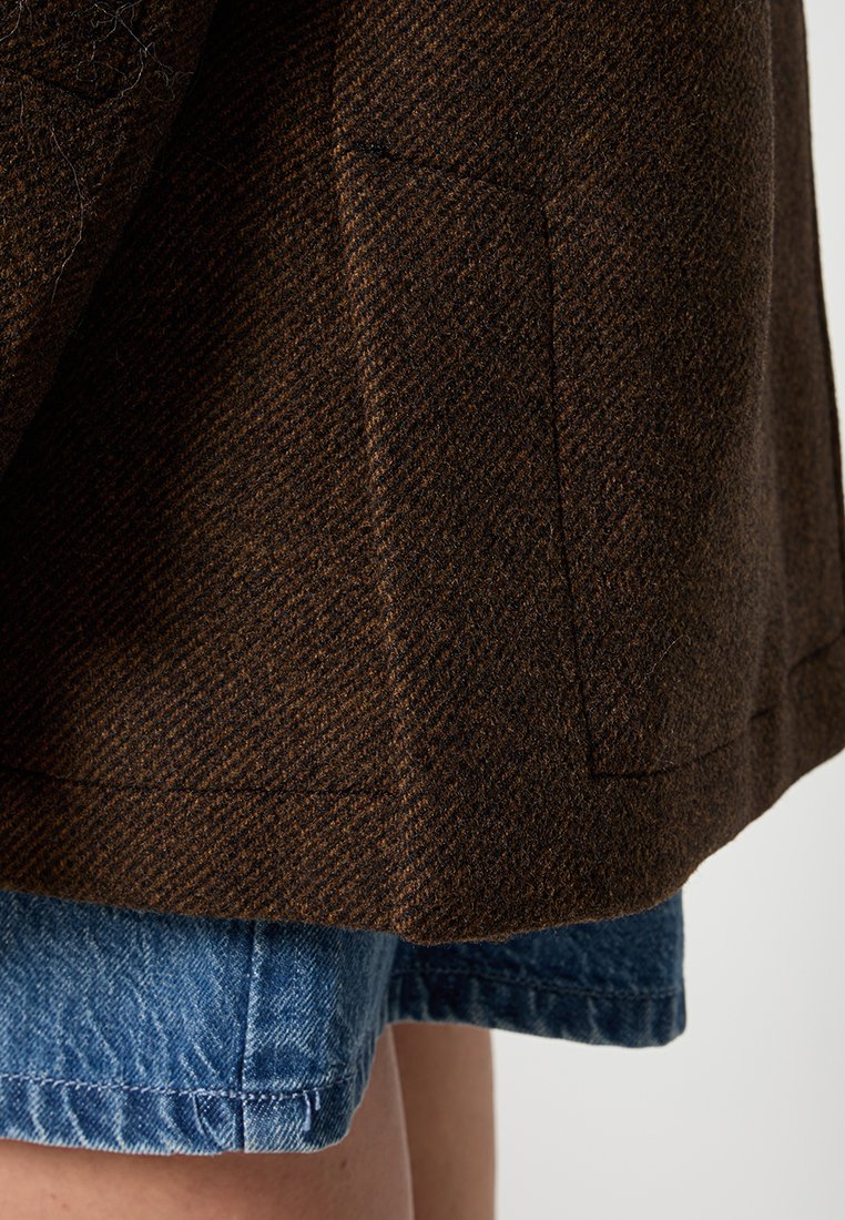 Bruine wollen jas met zichtbare zakstiksels, gedragen over blauwe denim shorts op een lichte huid.