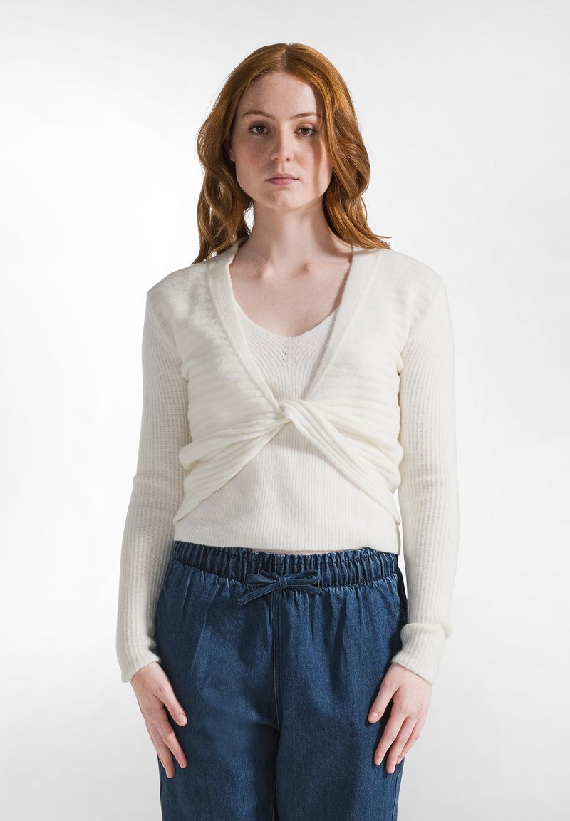 Maglione avvolgente bianco a maglia con maniche lunghe e scollo a V, abbinato a pantaloni in denim blu larghi con vita elasticizzata e dettaglio annodato.