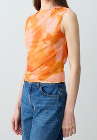 Femme portant un haut sans manches tie-dye orange et rose associé à un jean en denim bleu taille haute sur fond uni.