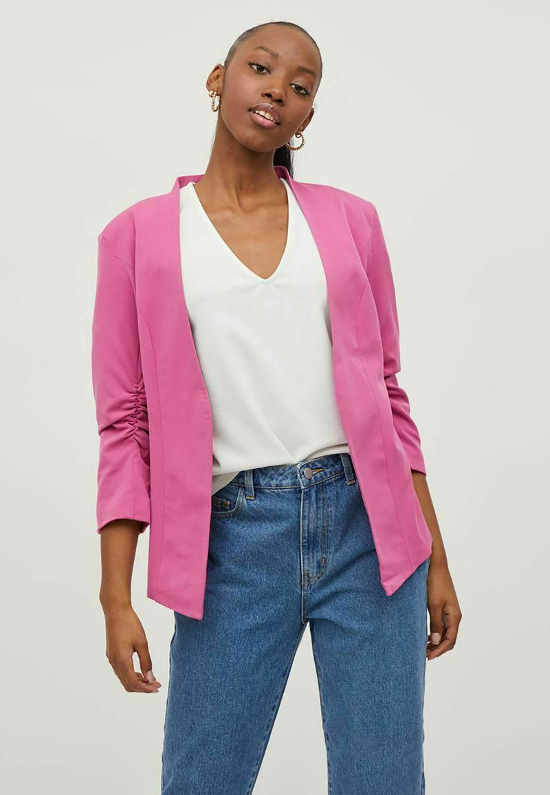 Rosa blazer med sjal-krage og trekvartlange ermer, kombinert med en hvit V-hals topp og blå høytlivsjeans.