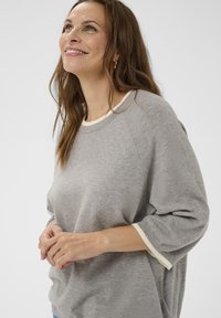 Nasmejana ženska oseba z dolgimi rjavimi lasmi, oblečena v sivo pulover s belim obrobo, gleda navzgor, roke sproščeno spočite na pasu, na beli podlagi.