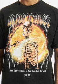 Sort bomulds t-shirt med grafik af et skelet mod en ildfarvet orange sol, med lynaccentuer og fed tekst ovenfor og nedenfor.