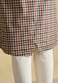 Zoom van het zoom van een geruit overhemd met een zijsplit over een witte broek, met een patroon van bruin, marineblauw en rood in het overhemd.