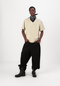 Chemise beige à manches courtes, pantalons noirs amples et bottines noires. La tenue comprend un bandana noir avec un motif blanc autour du cou.