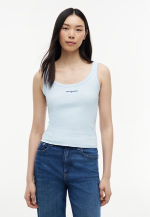 Młoda kobieta nosząca jasnoniebieski bezrękawnik z logo "tommy jeans" i niebieskie dżinsy z wysokim stanem, stojąca na białym tle.
