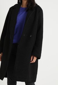 Černý, texturovaný kabát s rovnými liniemi. Obsahuje límec a dva přední knoflíky, nošený přes modrý svetr a černé kalhoty.