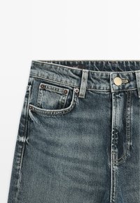 Džíny z denimu s tmavým odstínem, kontrastním prošitím, knoflíkovým zapínáním a přední mincovní kapsou. Textura je hladká s mírným vyblednutím.