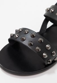 Sandalia negra de cuero con dos correas anchas adornadas con tachuelas metálicas. Suela negra lisa con un diseño minimalista.