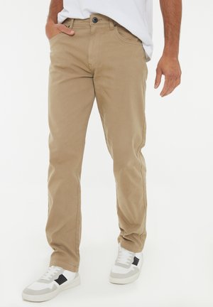 Pantalones de mezcla de algodón beige con un corte recto, bolsillos frontales y cierre de botón. Combinados con zapatillas blancas con acentos en negro.
