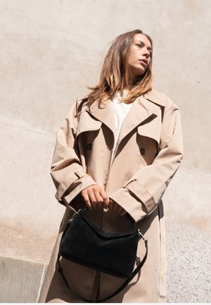 Femme en trench-coat beige tenant un sac à main noir contre un mur clair texturé, regardant sur le côté à la lumière naturelle.