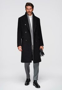 Abrigo negro de lana con doble botonadura y bolsillos laterales, llevado sobre un suéter de cuello alto blanco. Una bufanda de cuadros grises y guantes de cuero negros completan el look.