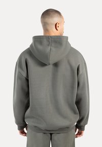 Homme avec les cheveux courts portant un sweat à capuche gris surdimensionné et un pantalon assorti, de dos contre un fond clair uni.