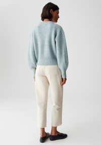 Femme debout de dos, portant un pull en laine côtelée bleu clair, un jean coupe crop blanc cassé avec des bords bruts, et des chaussures noires à enfiler.