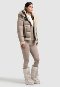 Beige Pufferjacke mit Lammfellkragen und -manschetten, kombiniert mit eng anliegenden beigen Leggings und weißen, mit Fell gefütterten Stiefeln.