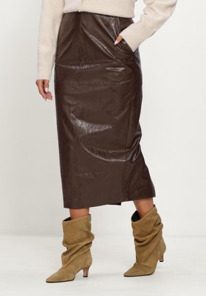 Vrouw met een glanzende donkerbruine leren midi-rok, beige losse suède enkellaarzen, en een crèmekleurige trui, staand met één hand in de zak.
