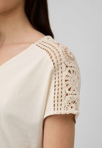 Chemise couleur crème avec un col en V et des détails en dentelle crochetée sur les épaules, ajoutant de la texture et des motifs délicats au design.