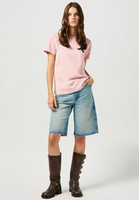 T-shirt in cotone rosa, vestibilità rilassata, con un piccolo logo. Pantaloni corti in denim azzurro chiaro con orli sfilacciati. Stivali marroni fino al ginocchio con fibbie.