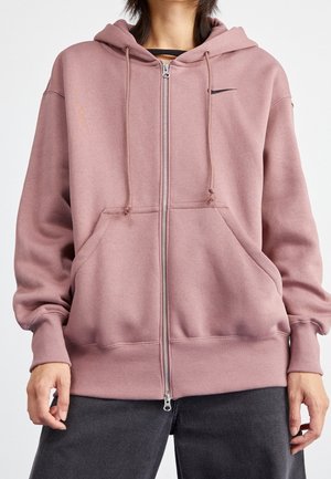 Personne portant un sweat à capuche mauve avec fermeture éclair, poches avant et cordons, associée à un pantalon gris foncé, sur un fond blanc uni.