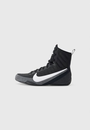 Czarny sportowy but Nike za kostkę z białym logo Swoosh, sznurowadłami i fakturowaną podeszwą, pokazany z profilu na jasnoszarym tle.