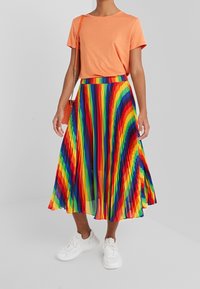 Jupe en arc-en-ciel plissée avec des rayures vibrantes, associée à un haut orange à manches courtes et des baskets blanches, avec un sac bandoulière.