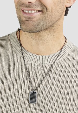 Pendentif en forme de plaque militaire en métal sur une fine chaîne, présentant un motif à carreaux. Porté sur un pull beige clair texturé avec un col côtelé.