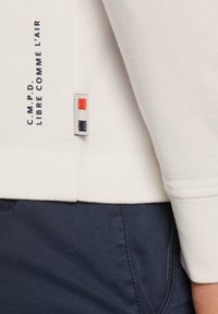 White long-sleeve top featuring a textured fabric, a knitted hem, and a tag with red and blue accents near the side seam.