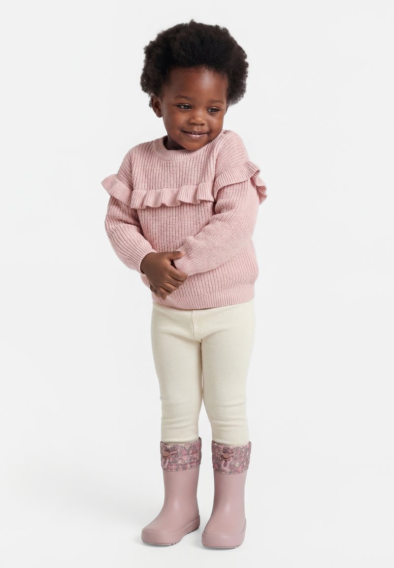 Petite fille portant un pull rose à volants, des leggings crème et des bottes roses avec des poignets floraux, debout, les mains jointes et souriante.