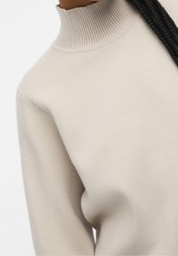 Beige gebreide trui met een hoge geribbelde kraag en een gladde textuur, met loszittende mouwen en een minimalistisch ontwerp.