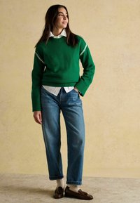 Grüner Strickpullover mit weißem Akzentmuster, kombiniert mit hellblauen geraden Jeans und braunen Slippers auf weißen Socken. Versteckte Knopfhülle.