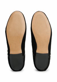 Par de mocasines negros mostrados desde la vista de la suela, con suelas de cuero marrón claro y agarres de goma negra en el tacón.