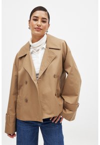Karamellfärgad dubbelknäppt jacka med breda slagkragar, knappdetaljer och manschettremmar. Bärs över en vit ribbad polotröja och denim.
