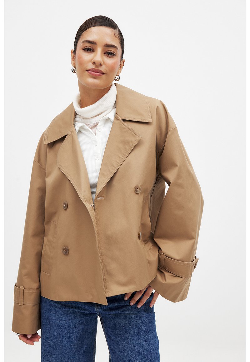 Karamellfärgad dubbelknäppt jacka med breda slagkragar, knappdetaljer och manschettremmar. Bärs över en vit ribbad polotröja och denim.