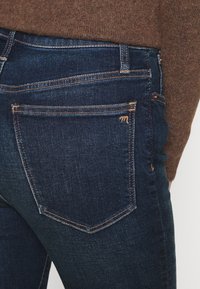 Primer plano de un bolsillo trasero de jeans de mezclilla azul oscuro con costura naranja, combinado con un suéter marrón.