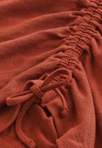 Tissu côtelé couleur rouille avec taille élastiquée froncée et détail de cordon. Texture douce avec une finition lisse et un léger éclat.