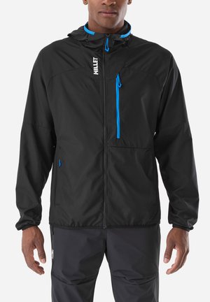 Homme portant une veste légère noire avec des accents de fermeture éclair bleus et une capuche, debout les bras détendus le long du corps.