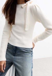 Lekki kremowy sweter dziany z bufkami na rękawach i wiązaniem przy dekolcie, zestawiony z niebieskimi dżinsami. Miękka tekstura.