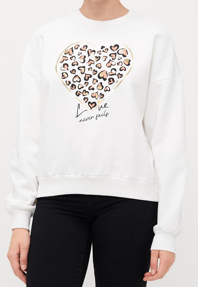 Sweat-shirt blanc avec un grand cœur composé de cœurs imprimés léopard et le texte « L’amour ne faillit jamais », porté avec un pantalon noir.