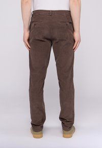 Hnědé kordové kalhoty s propracovaným střihem, dvěma zadními kapsami a jemnou texturou. Nohy se mírně zužují směrem k lemu.