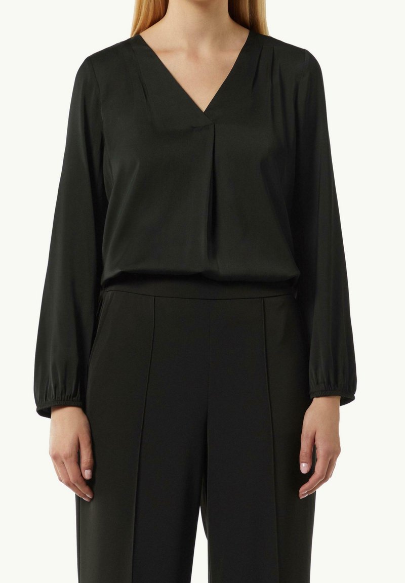 Femme portant un chemisier noir à manches longues et col en V, rentré dans un pantalon noir taille haute avec des détails de coutures visibles.