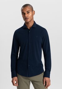 Navyblaues Langarm-Hemd mit Knöpfen, aus weichem Stoff, mit klassischem Kragen und subtilen Logodetails. Abgerundet durch einen geschwungenen Saum.