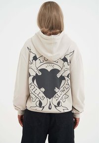 Beżowa bluza z kapturem z dużym czarnym wzorem graficznym przedstawiającym serce, czaszki oraz napis "MIŁOŚĆ ZABIJA POWOLI" na plecach.