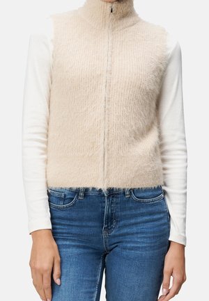 Chaleco sin mangas beige y peludo con cremallera frontal, usado sobre una camisa blanca de manga larga, combinado con jeans de mezclilla azul.