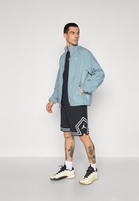 Chaqueta azul clara con un cuello alto, cierre de cremallera y patrón de puntos; camiseta negra, pantalones cortos de malla negra con acentos blancos; zapatillas deportivas color crema.
