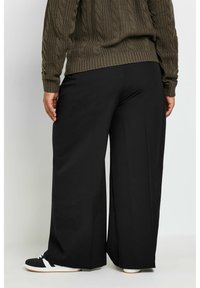 Schwarze, weit geschnittene Hosen aus weichem Material mit glatter Textur und passgenauer Passform, kombiniert mit einem dunkelgrünen, strukturierten Strickpullover.
